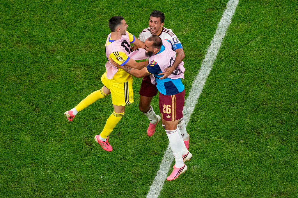 Three soccer players celebrate a goal, jumping and embracing on a green field near a white sideline line in mid-air.
