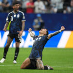 Soccer player in a blue kit kneels on the field, arms raised in celebration as teammates and spectators look on.
