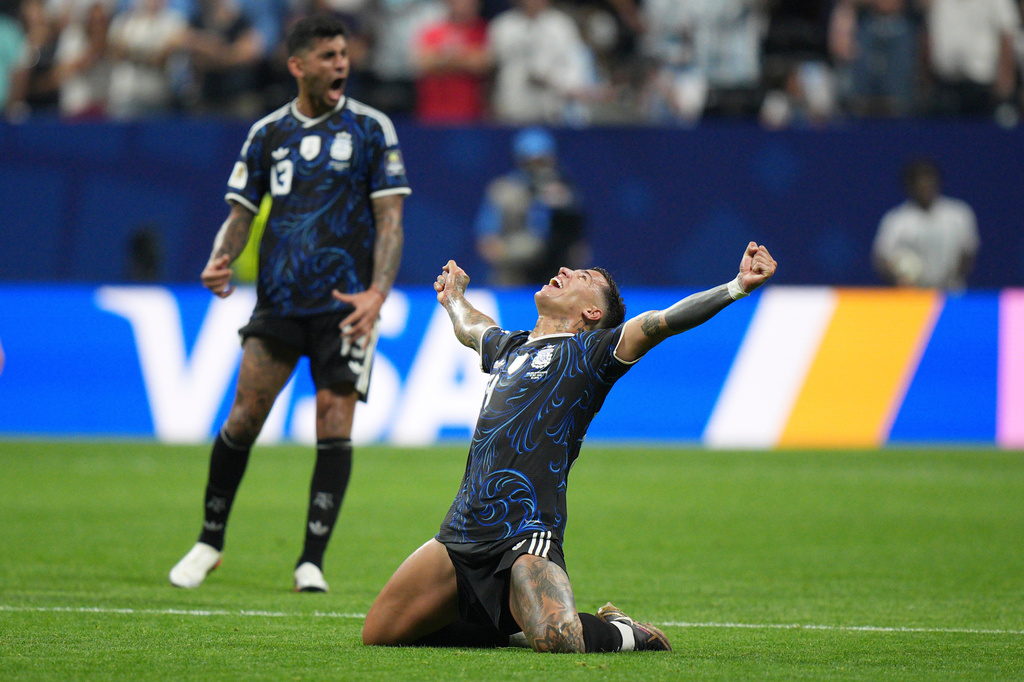 Soccer player in a blue kit kneels on the field, arms raised in celebration as teammates and spectators look on.