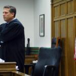 A judge in a black robe stands with arms crossed in a courtroom beside a U.S. flag and dark wood paneling.