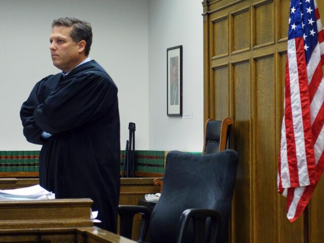 A judge in a black robe stands with arms crossed in a courtroom beside a U.S. flag and dark wood paneling.