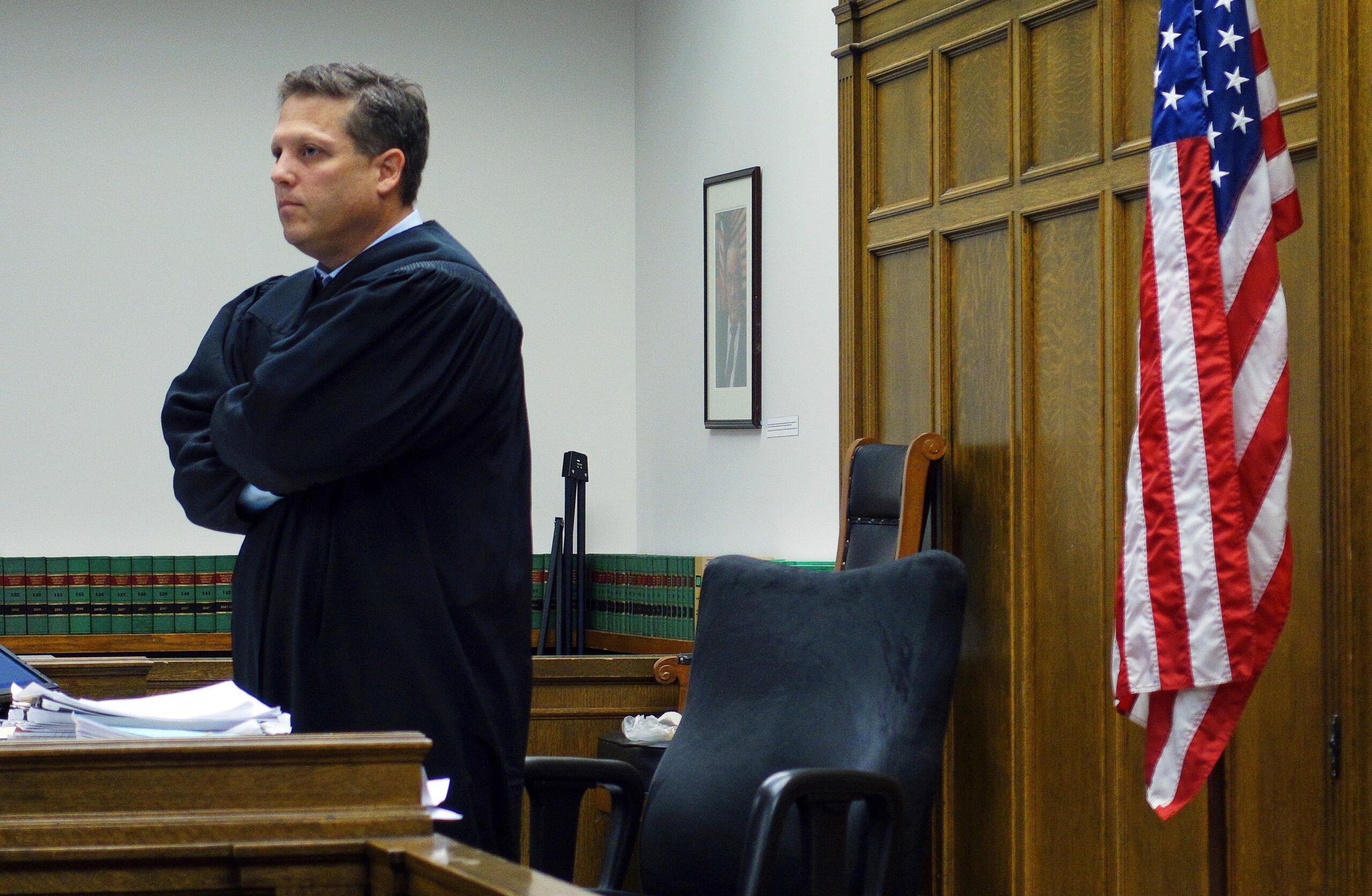 A judge in a black robe stands with arms crossed in a courtroom beside a U.S. flag and dark wood paneling.