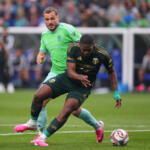 Two soccer players battle for the ball during a match on a green field, with one in a bright green jersey and the other in dark kit close in pursuit.