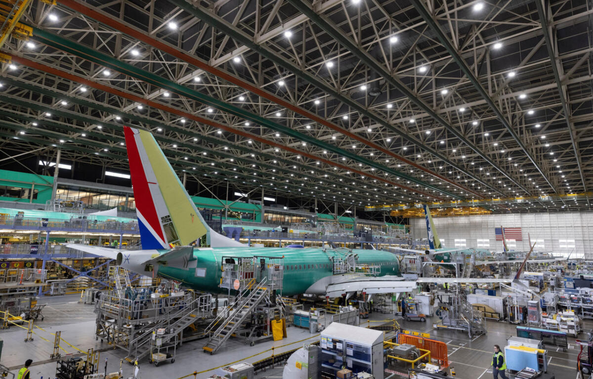 Inside a large aircraft hangar, a green fuselage airliner is on a mobile platform with scaffolding and workers nearby