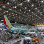 Inside a large aircraft hangar, a green fuselage airliner is on a mobile platform with scaffolding and workers nearby
