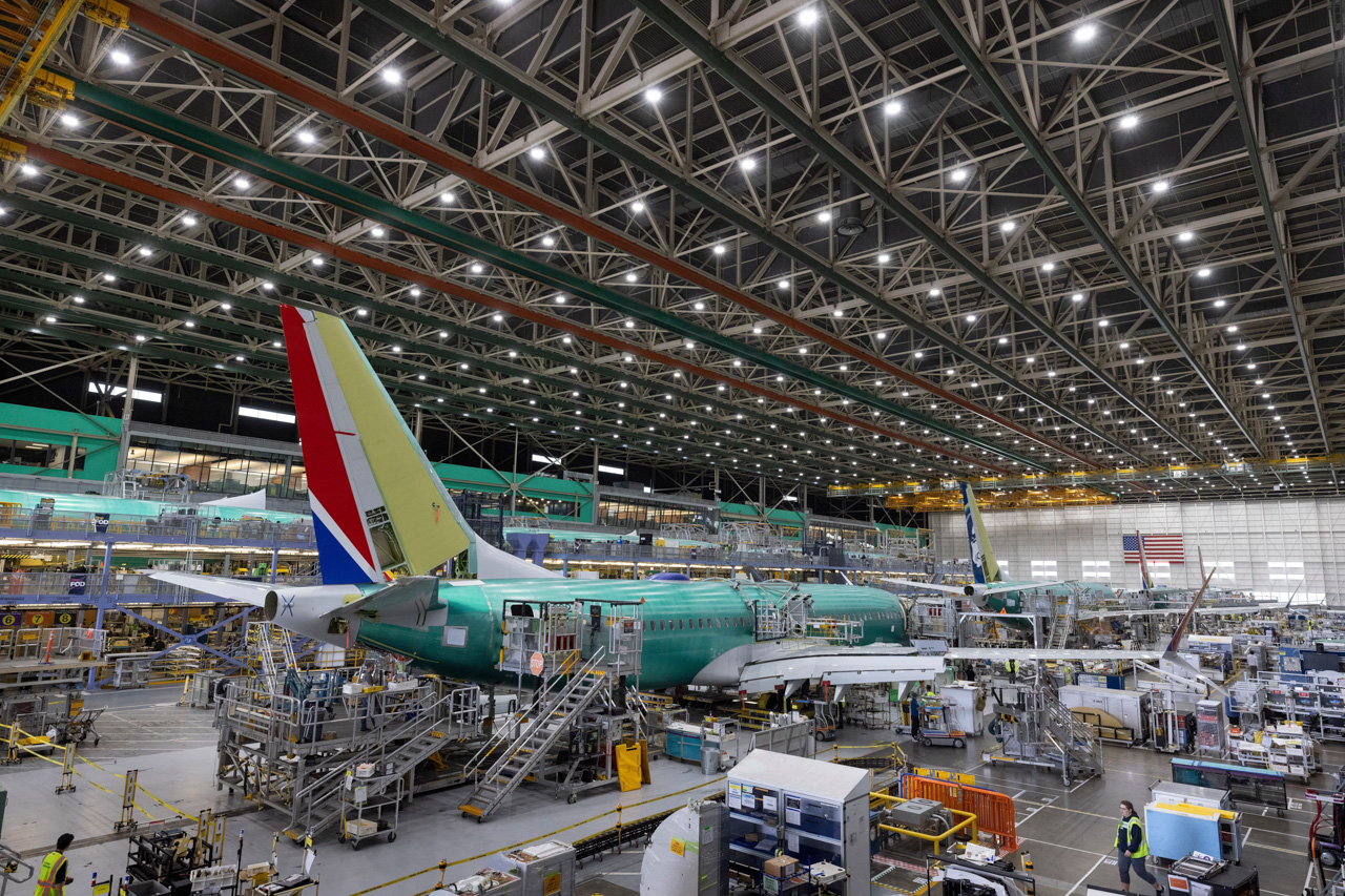 Inside a large aircraft hangar, a green fuselage airliner is on a mobile platform with scaffolding and workers nearby