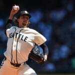 Pitcher in a Seattle Mariners uniform mid-throw, ball raised ready to pitch on a sunny field with a blurred crowd in the background