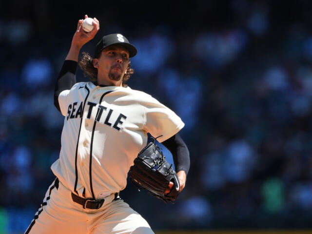 Pitcher in a Seattle Mariners uniform mid-throw, ball raised ready to pitch on a sunny field with a blurred crowd in the background