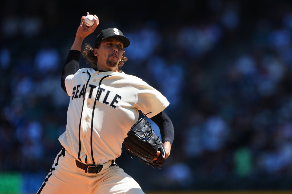 Pitcher in a Seattle Mariners uniform mid-throw, ball raised ready to pitch on a sunny field with a blurred crowd in the background