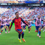 Spanish soccer player in a red jersey (number 7) runs toward the camera as teammates in pink bibs chase on a green field during a World Cup match with a packed stadium in the background.