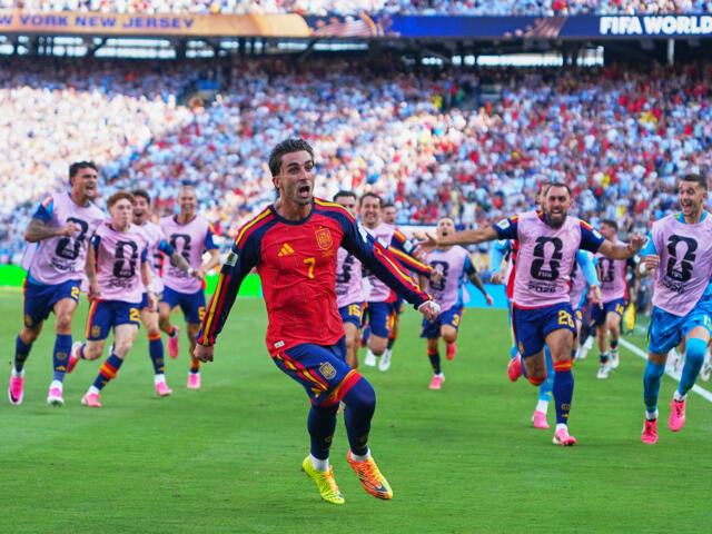 Spanish soccer player in a red jersey (number 7) runs toward the camera as teammates in pink bibs chase on a green field during a World Cup match with a packed stadium in the background.