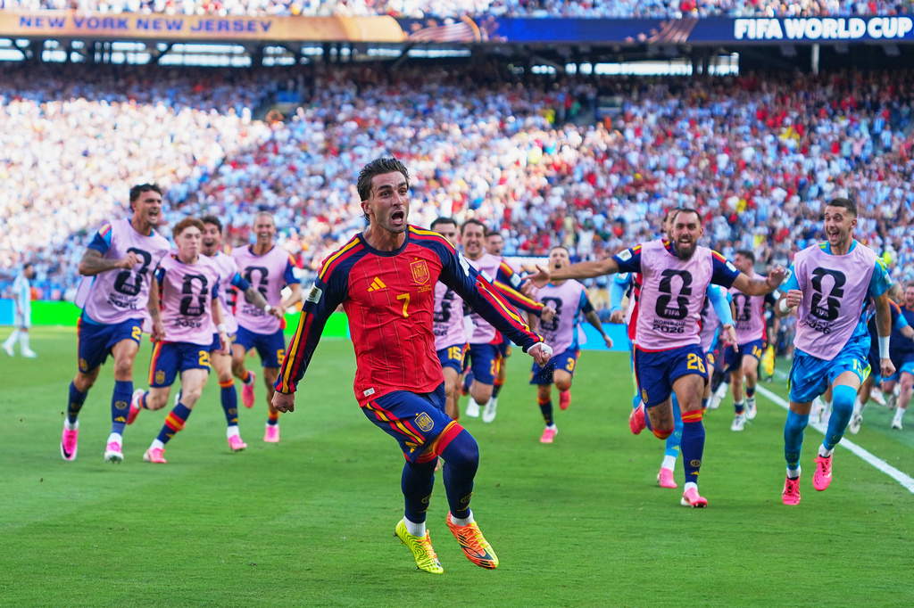 Spanish soccer player in a red jersey (number 7) runs toward the camera as teammates in pink bibs chase on a green field during a World Cup match with a packed stadium in the background.