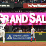 Baseball player in a white uniform with number 29 raises his arm toward a large pink 'GRAND SLAM' banner as a gray-uniform opponent stands nearby on the field.