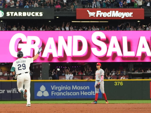 Baseball player in a white uniform with number 29 raises his arm toward a large pink 'GRAND SLAM' banner as a gray-uniform opponent stands nearby on the field.