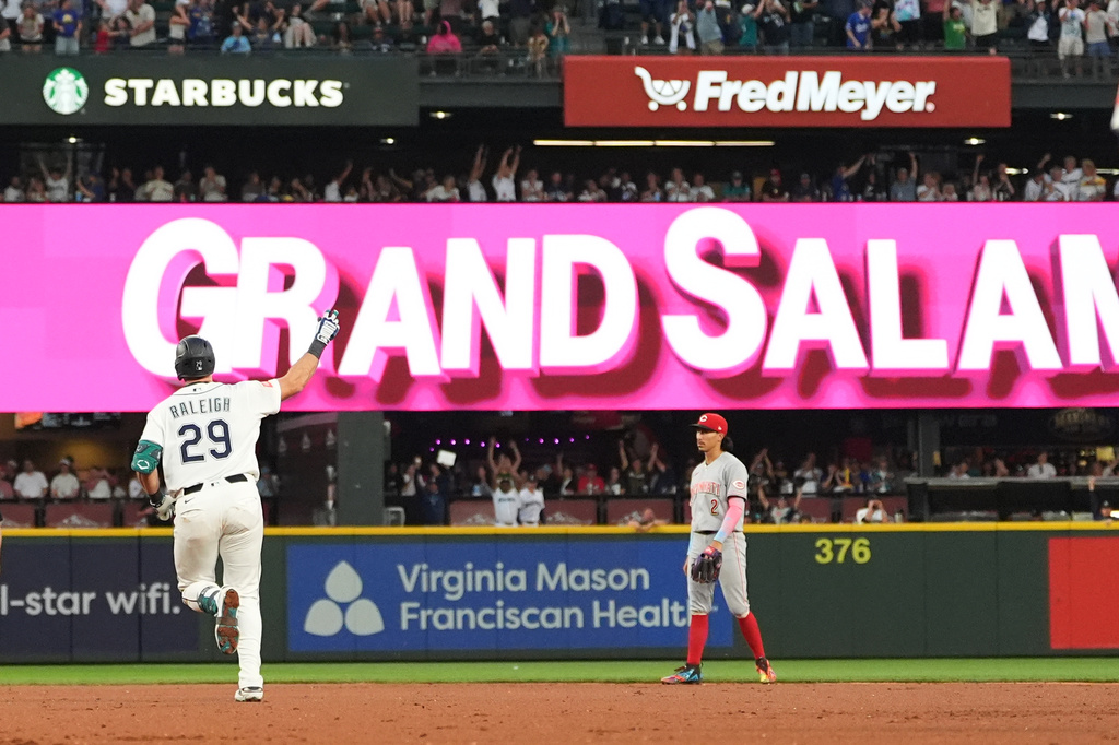 Baseball player in a white uniform with number 29 raises his arm toward a large pink 'GRAND SLAM' banner as a gray-uniform opponent stands nearby on the field.