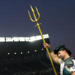 Baseball catcher raises a large gold trident during a game at dusk in a stadium park.