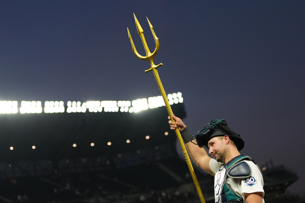 Baseball catcher raises a large gold trident during a game at dusk in a stadium park.