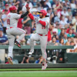 Two baseball players in gray uniforms high-five while jumping on the field as the crowd cheers behind them.