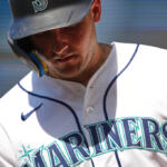 Close-up of a baseball player wearing a Mariners helmet and white jersey, looking down at the ground.