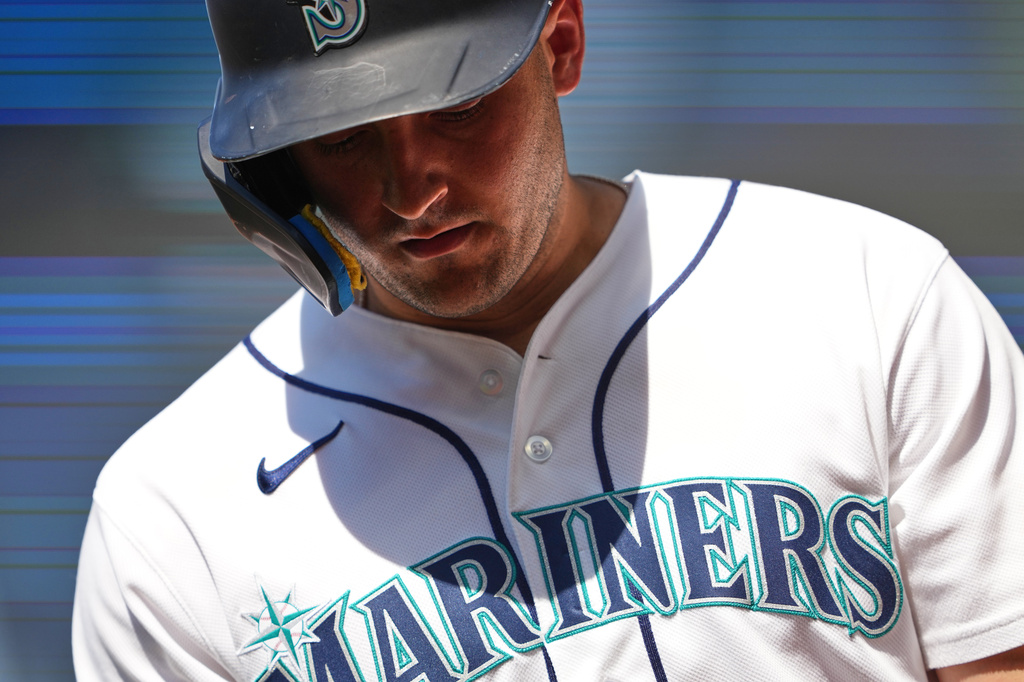Close-up of a baseball player wearing a Mariners helmet and white jersey, looking down at the ground.