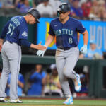 Two Seattle Mariners players in blue uniforms high-five on the baseball field during a game, one running toward the other with gloves on.