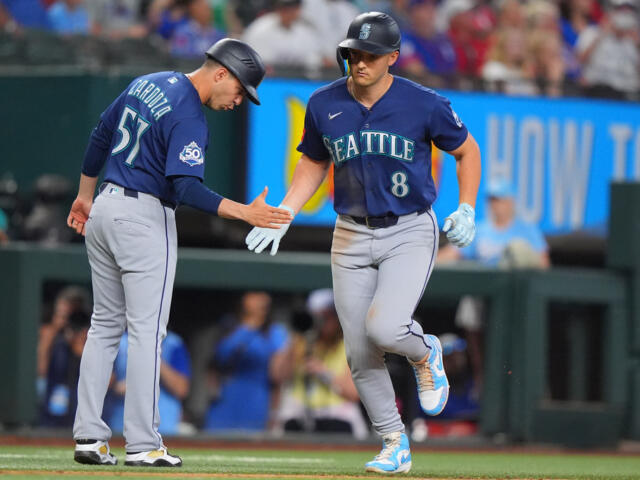 Two Seattle Mariners players in blue uniforms high-five on the baseball field during a game, one running toward the other with gloves on.