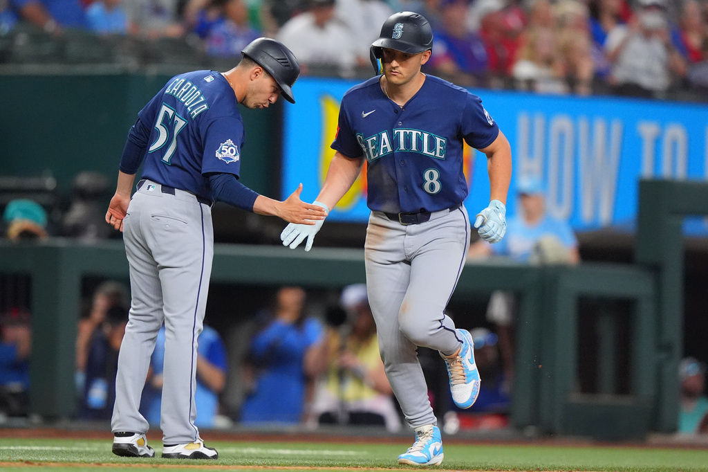 Two Seattle Mariners players in blue uniforms high-five on the baseball field during a game, one running toward the other with gloves on.