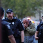 A distressed man with hands covering his face walks between two police officers in tactical vests.