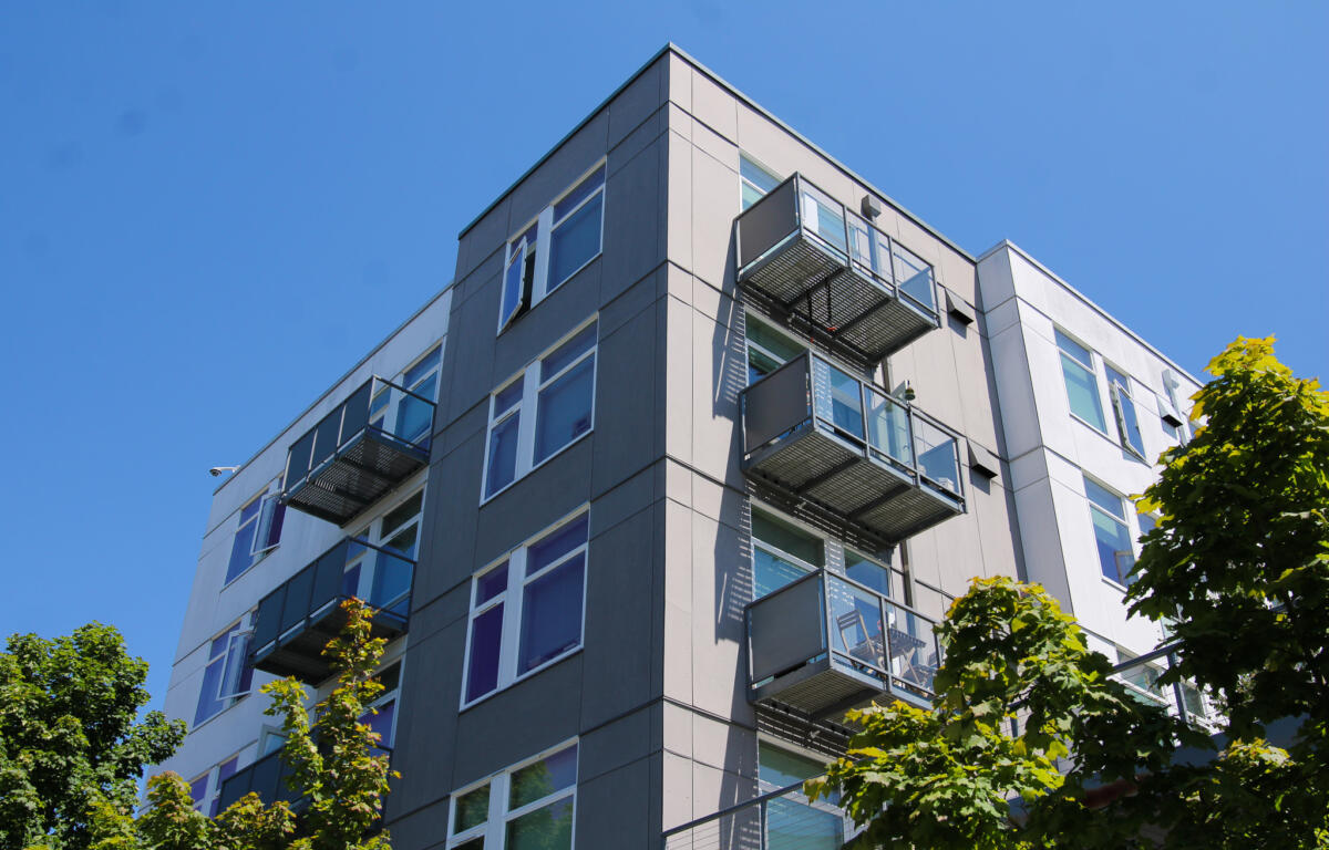Corner view of a modern gray apartment building with metal balconies set against a clear blue sky, with green trees in the foreground.