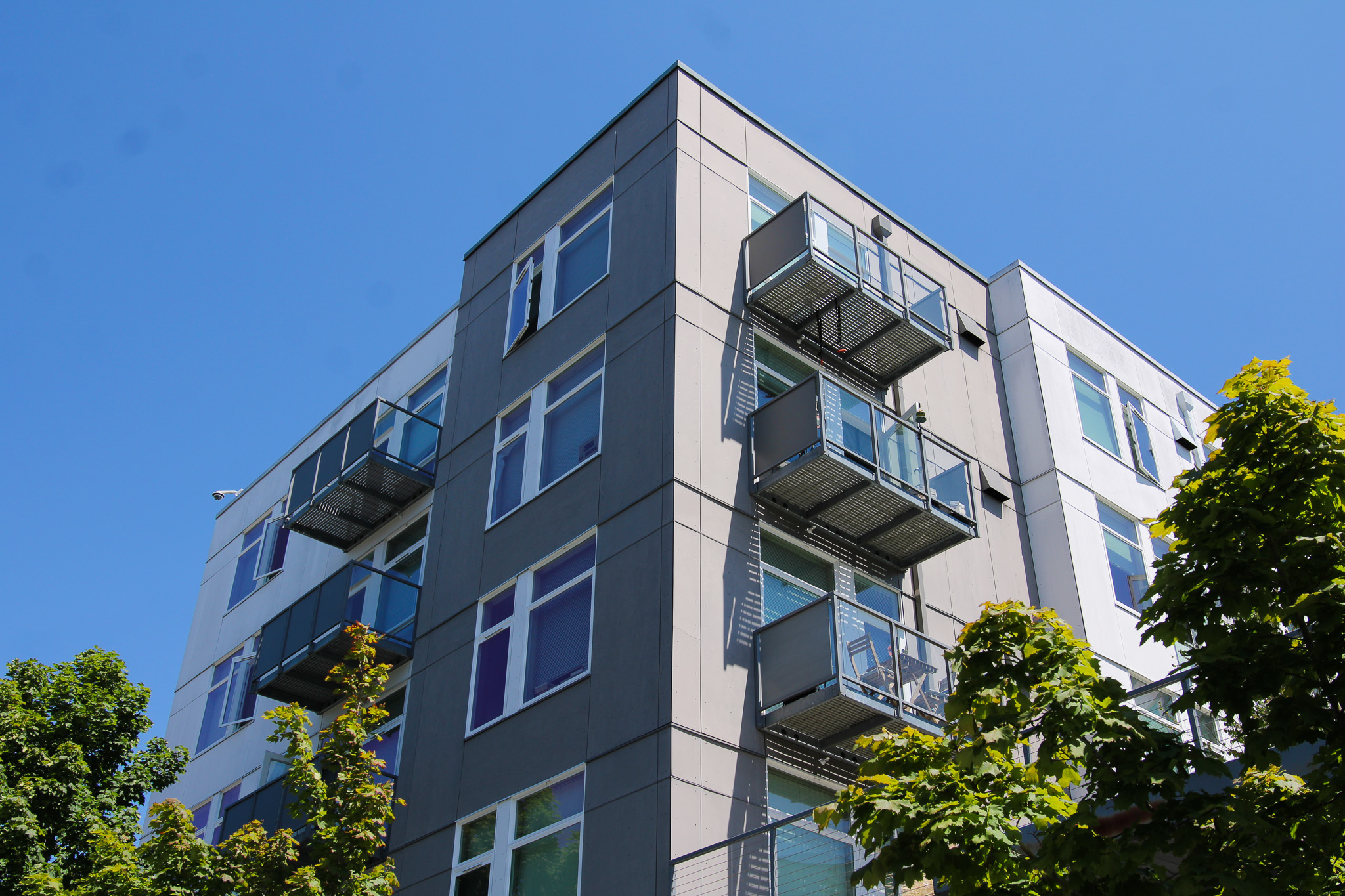Corner view of a modern gray apartment building with metal balconies set against a clear blue sky, with green trees in the foreground.