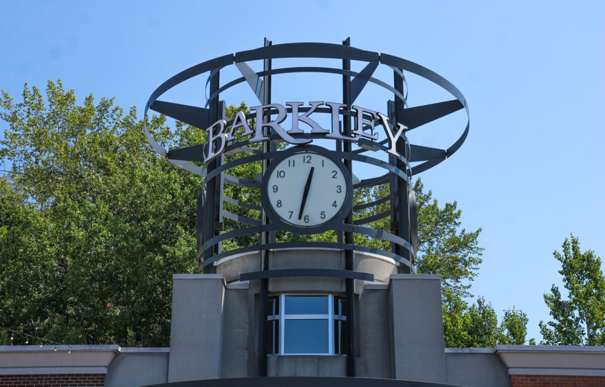 Clock tower structure with a round clock and 'BARKLEY' sign against a blue sky, trees in the background.