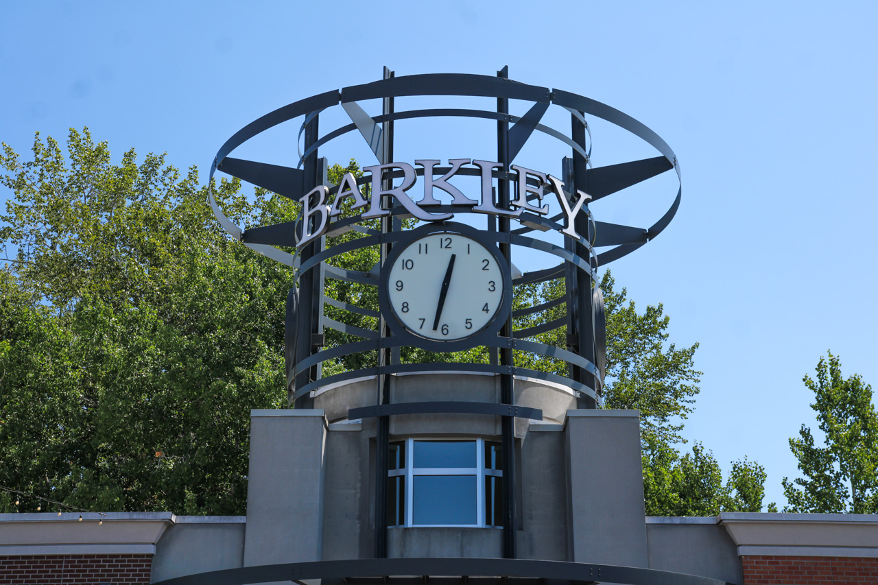 Clock tower structure with a round clock and 'BARKLEY' sign against a blue sky, trees in the background.