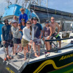 Group of ten people posing on the deck of a sailboat named ABSOLUTELY at a sunny marina.