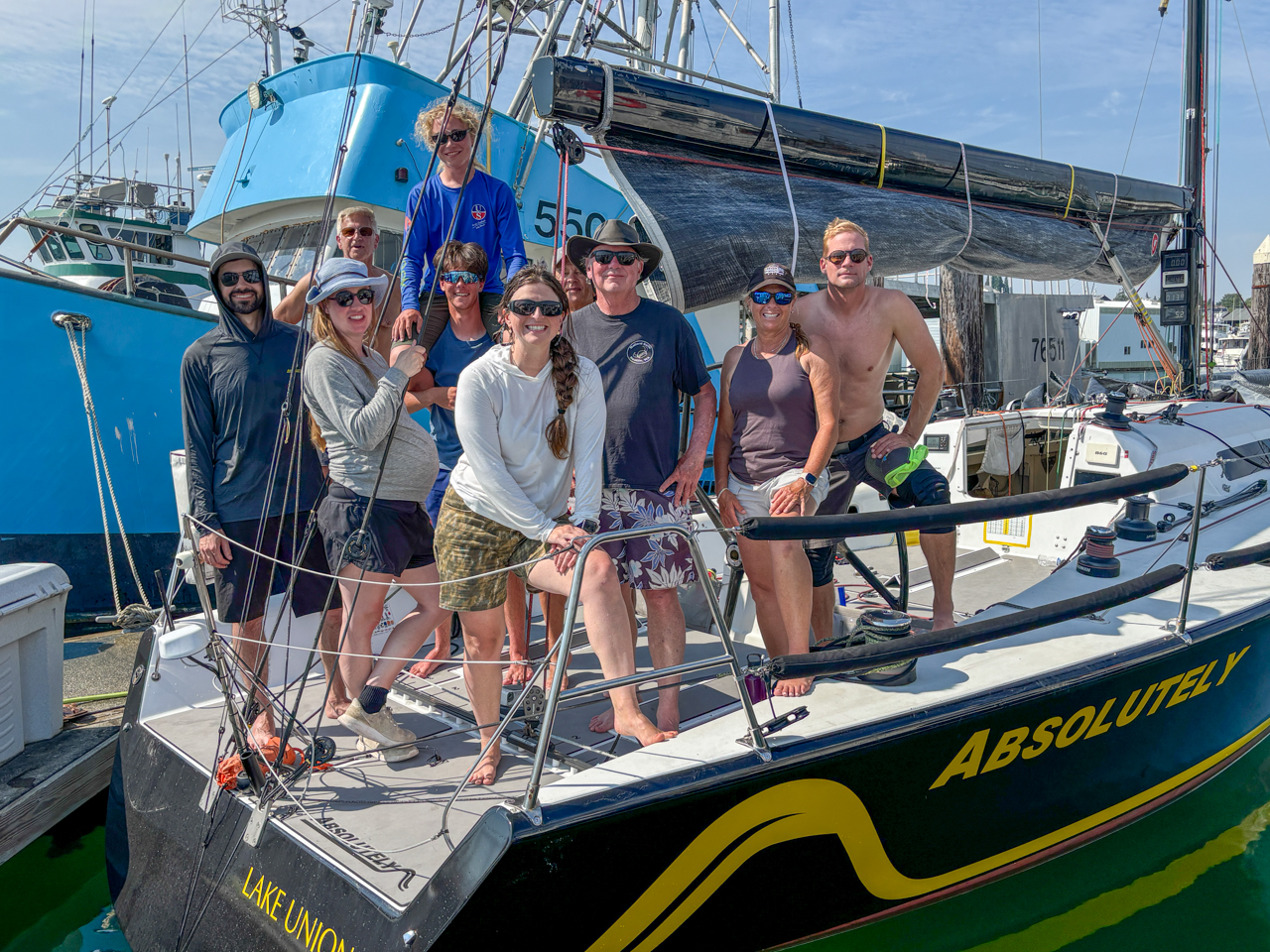 Group of ten people posing on the deck of a sailboat named ABSOLUTELY at a sunny marina.