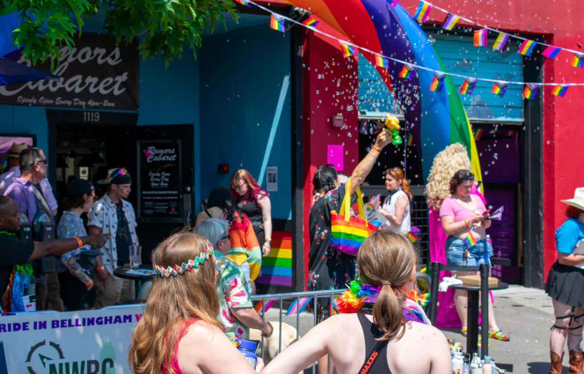 Rainbow Pride parade on a sunny street with people in colorful outfits and confetti in the air near rainbow banners and storefronts outside a club venue.