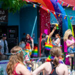 Rainbow Pride parade on a sunny street with people in colorful outfits and confetti in the air near rainbow banners and storefronts outside a club venue.