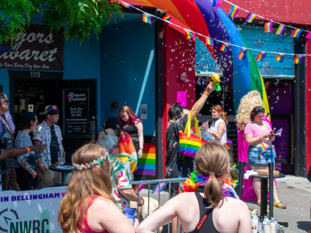 Rainbow Pride parade on a sunny street with people in colorful outfits and confetti in the air near rainbow banners and storefronts outside a club venue.