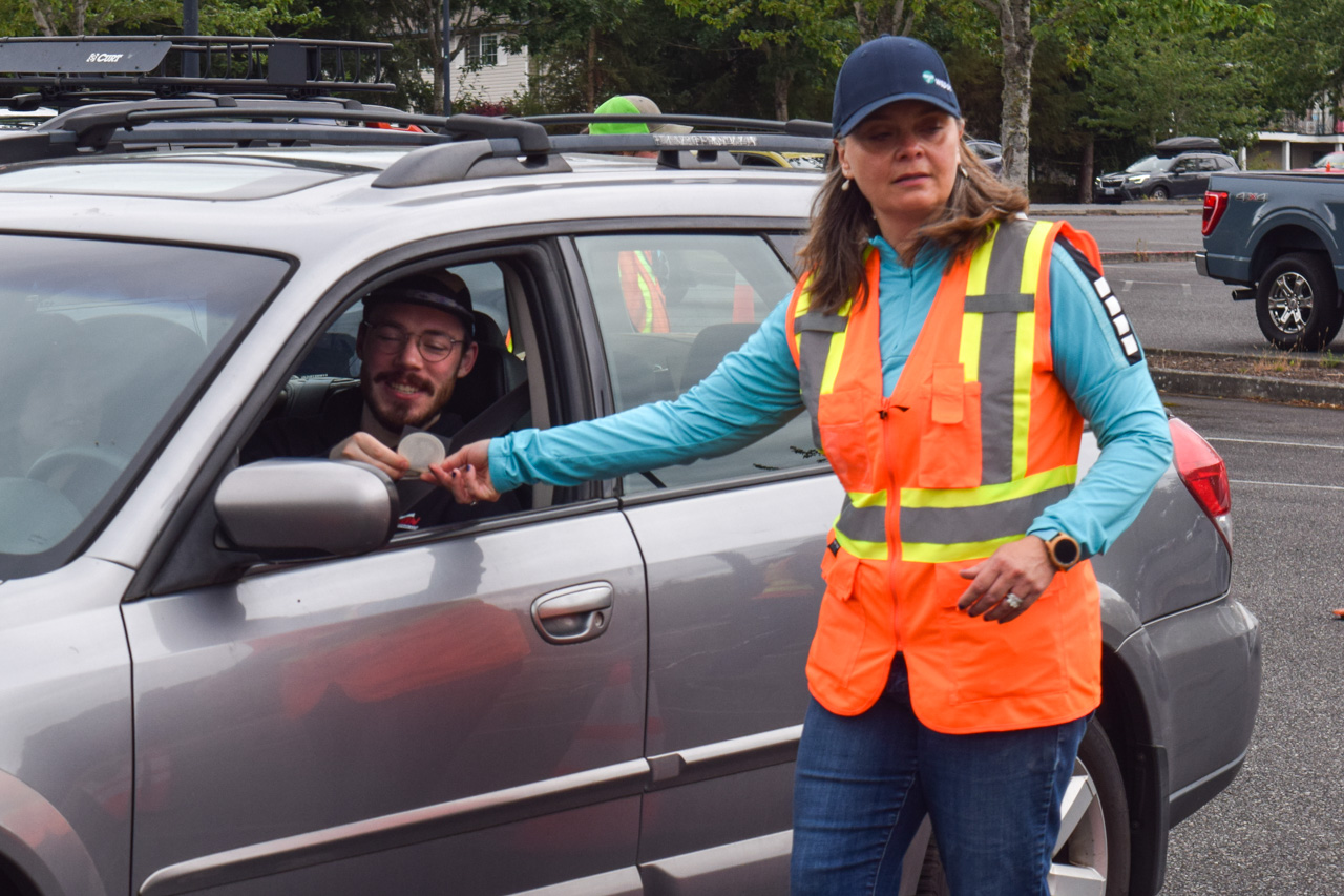 Safety worker in a bright orange reflective vest hands a round item to a smiling driver in a silver car window. RGB scene in a parking lot or checkpoint area: a woman extends an object toward the car window while wearing a cap and high-visibility vest.