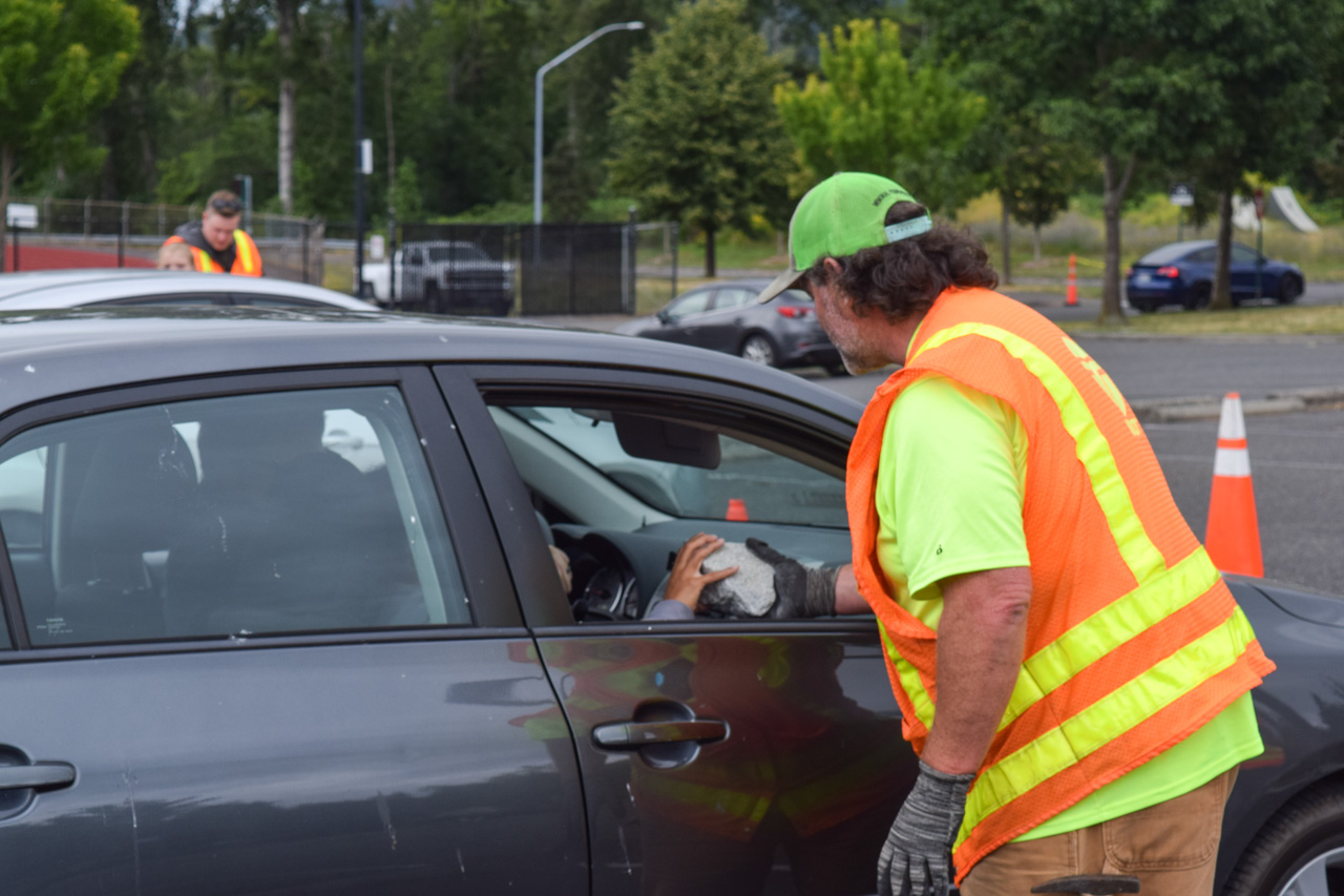 Worker in a high-visibility vest hands a rock to a driver through a car window at a roadside safety checkpoint; cones and trees in the background.