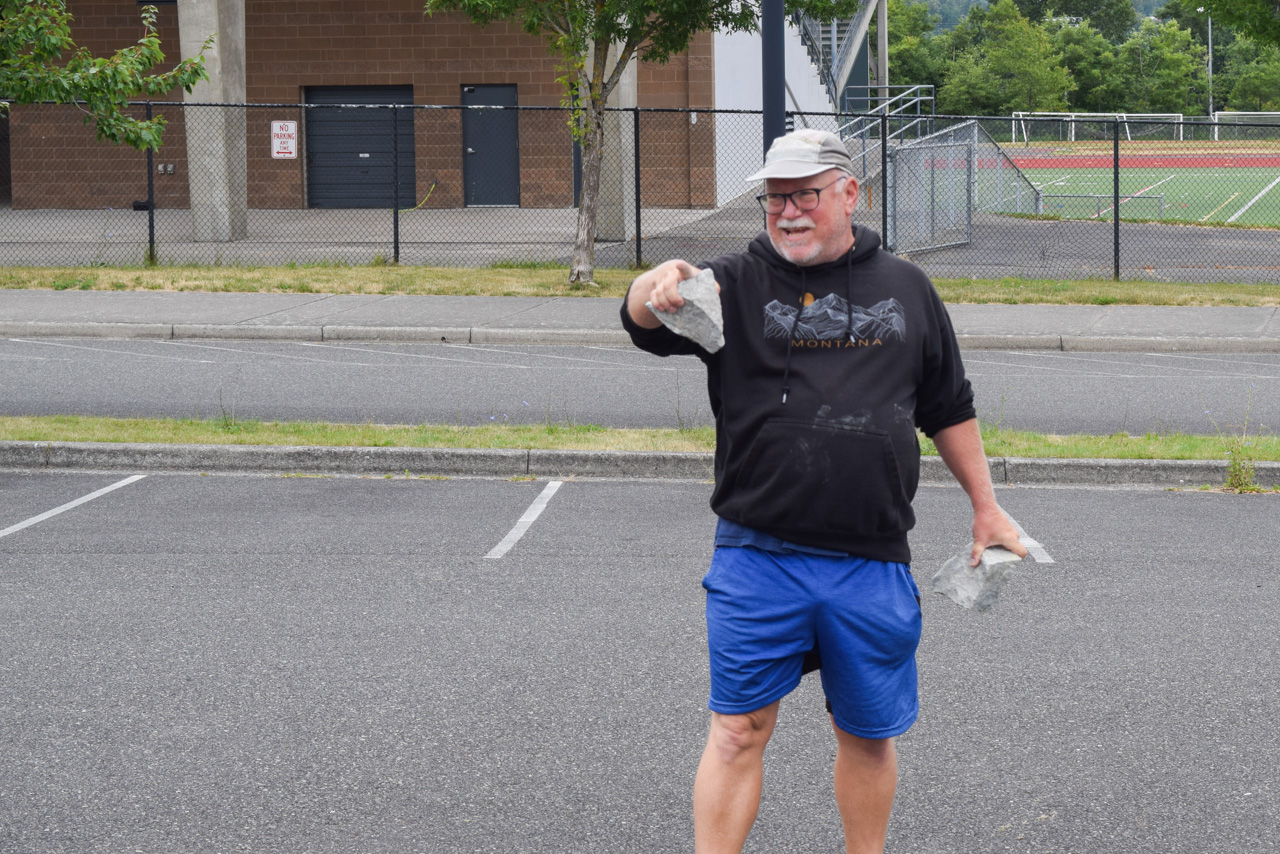 Older man in a Montana hoodie and blue shorts throws a gray rock in a parking lot; chain-link fence and sports field behind him.