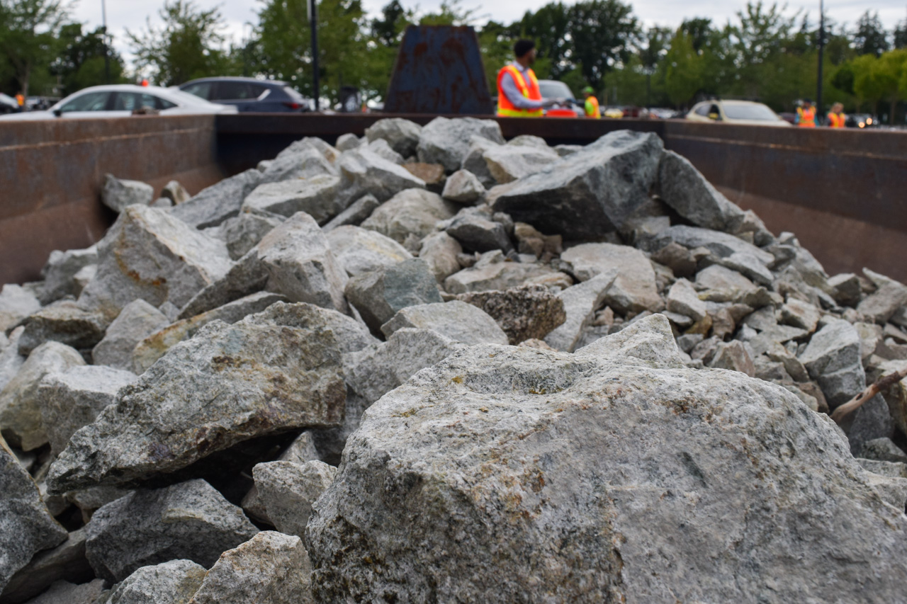 Pile of gray rocks and rubble in a truck bed, with workers in high-visibility vests in the background.