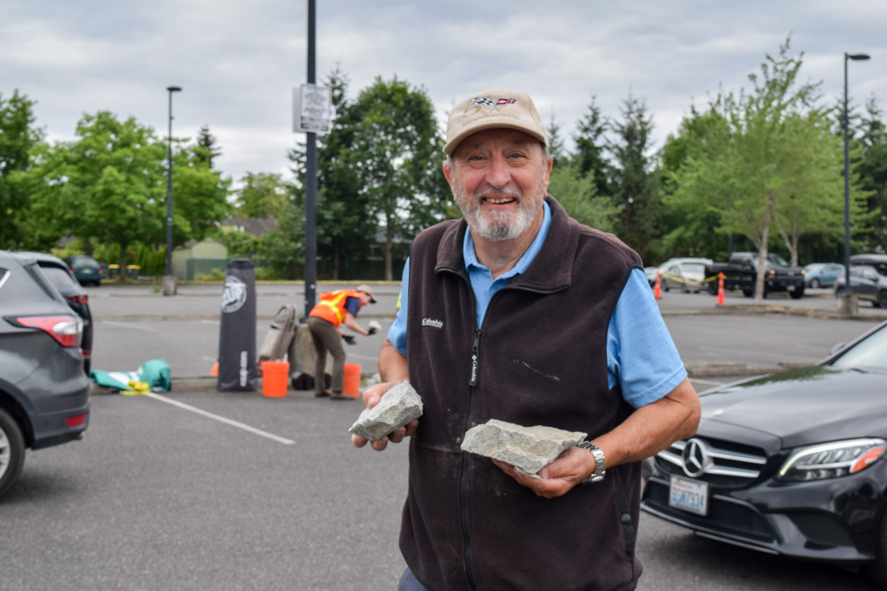 Smiling older man in a cap and vest holds two rocks in a parking lot during a cleanup event.