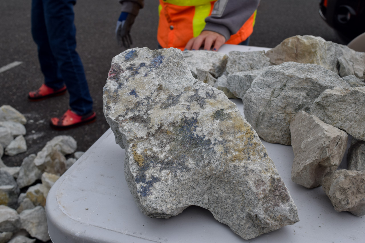 Rough, multi-colored rock resting on a table with construction workers in safety vests and jeans in the background, suggesting a geology or rock sample setting.