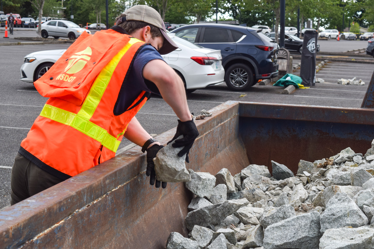 Utility worker in an orange safety vest and cap loads debris into a rusted dumpster in a parking lot.
