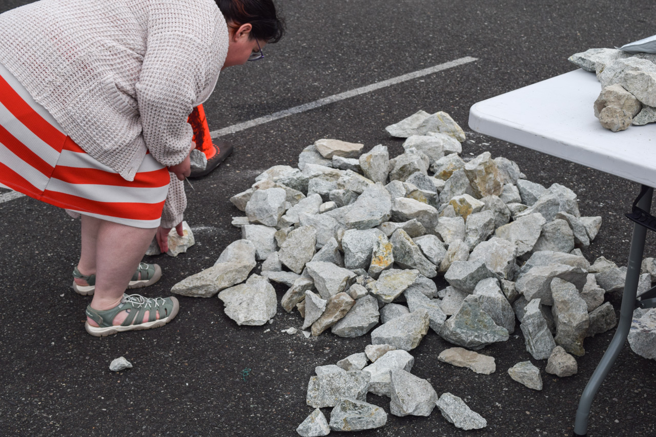 Person crouching to sort gray stones on asphalt next to a white folding table with rocks nearby