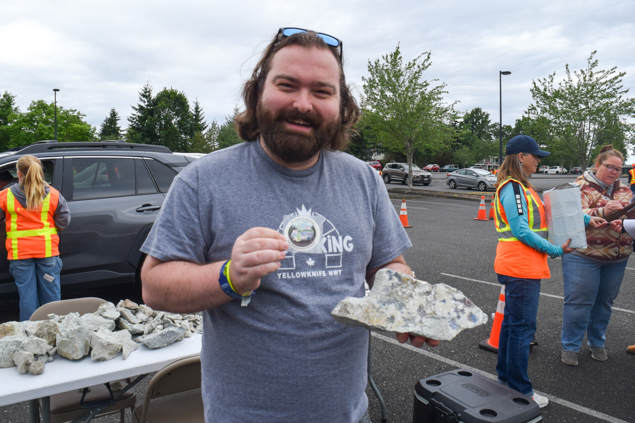 Smiling man in a gray t-shirt holding a large rock sample at an outdoor rock-collecting event, table of rocks in the foreground.