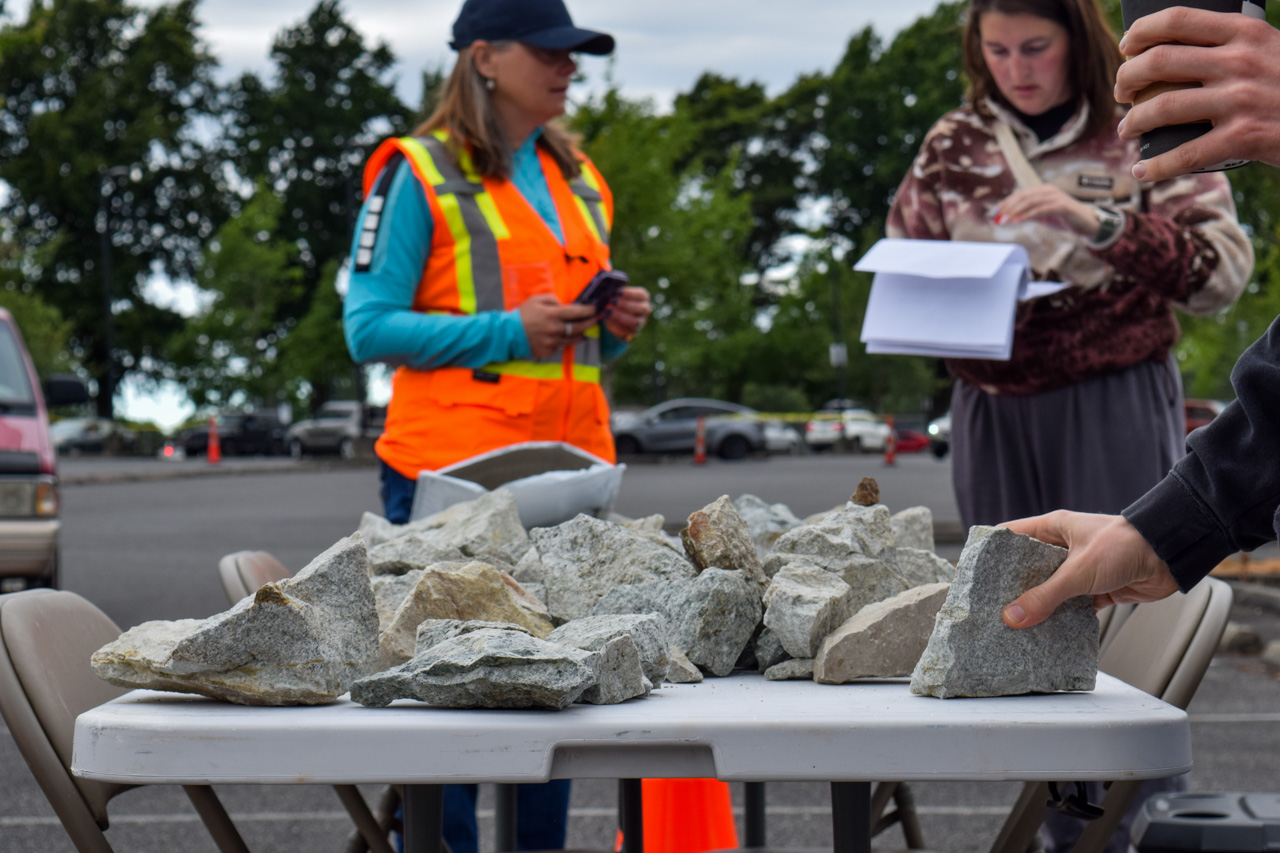 Rocks arranged on a white table at an outdoor parking-lot workshop; a person in a bright safety vest talks in the background.