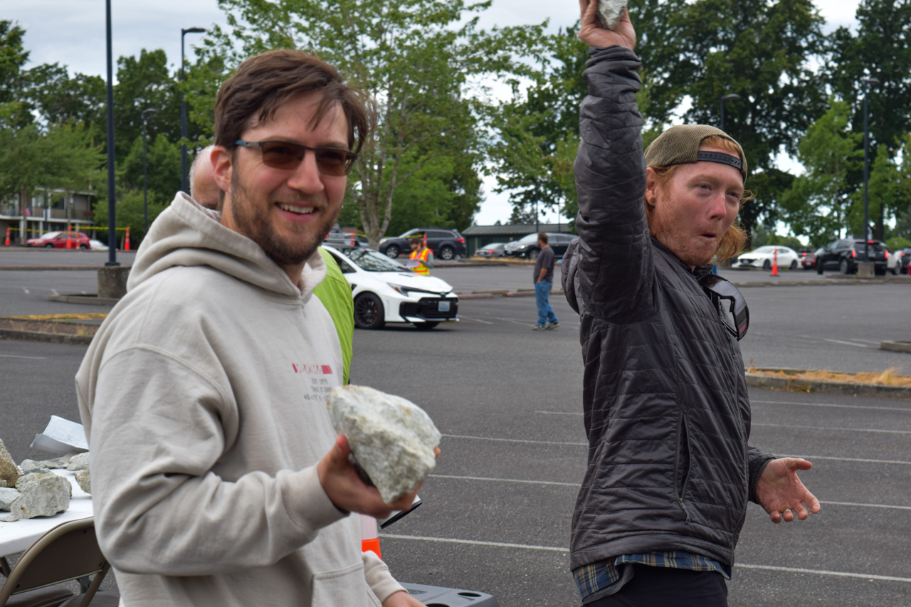 Two men in a parking lot, one smiling while holds a large rock, the other raises a rock overhead in a playful moment