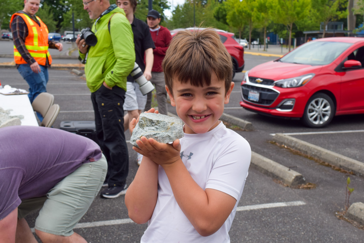 Smiling boy in a white shirt holds up a rock in a parking lot, with photographers and a red car in the background.
