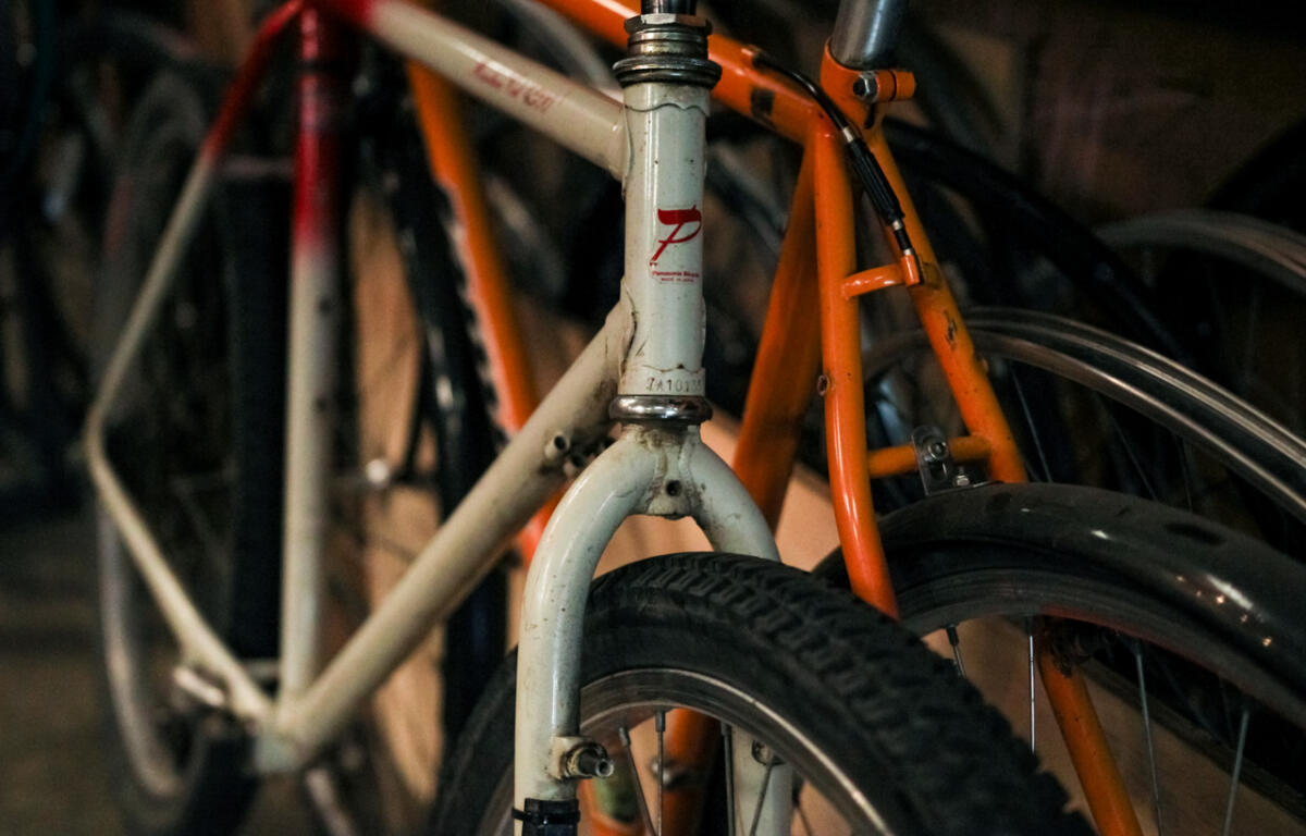 Close-up of a white vintage bicycle head tube and front fork with a red logo, with an orange bike in the background in dim storage.